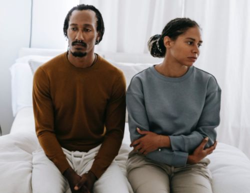 Illustration: Couple sitting side by side on a bed, looking in different directions with sad expressions.