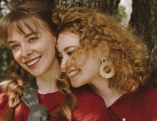 Illustration: Two women in red dresses standing close and laughing in front of a large tree trunk. 